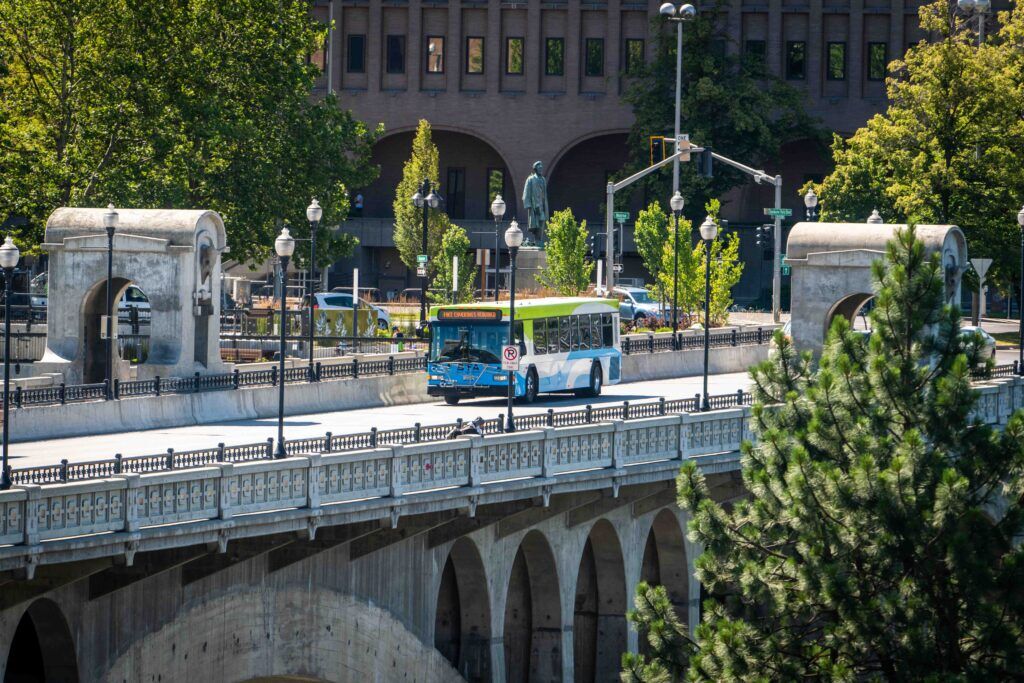 A blue and white public bus crosses an arched stone bridge on a sunny day. Trees and a large brick building with many windows are visible in the background. Streetlights and pedestrians can also be seen on and around the bridge.