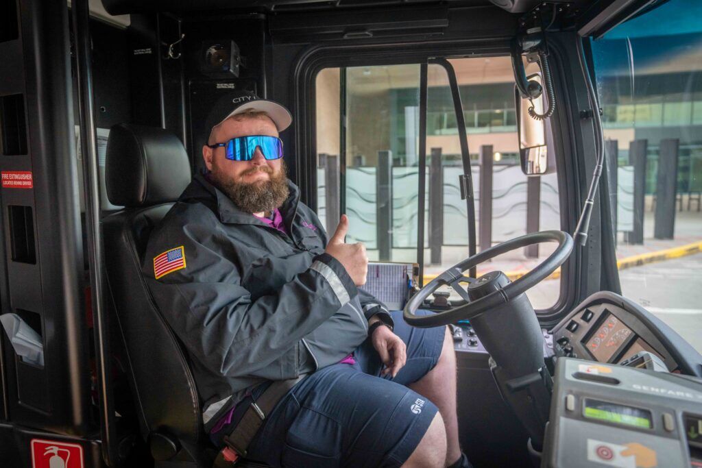 Bus driver giving a thumbs up in the driver's seat, wearing sunglasses and a jacket.