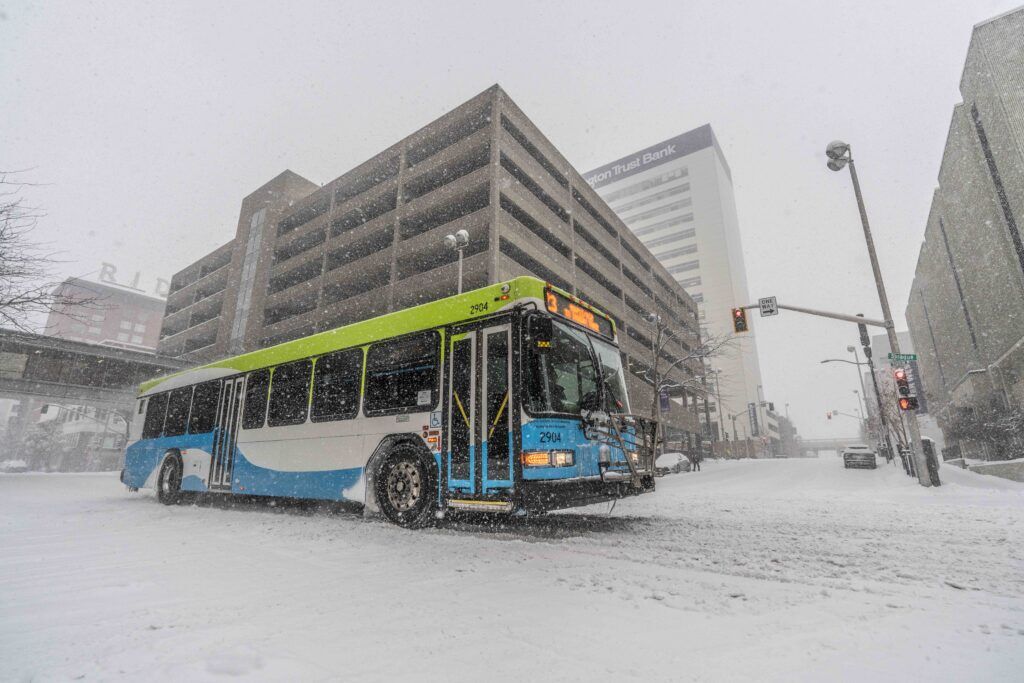 STA bus driving through heavy snowfall on a snowy Spokane street with buildings in the background.